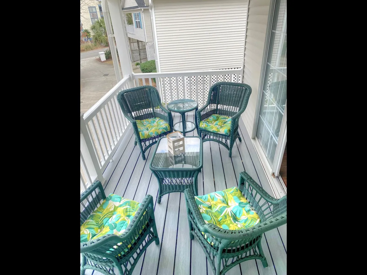 A casual and welcoming porch area with green wicker chairs, a small glass table, and colorful cushions. The space feels perfect for sunset views, enjoying the morning sunlight, or sharing stories with neighbors, highlighting the social possibilities of the cottage complex.