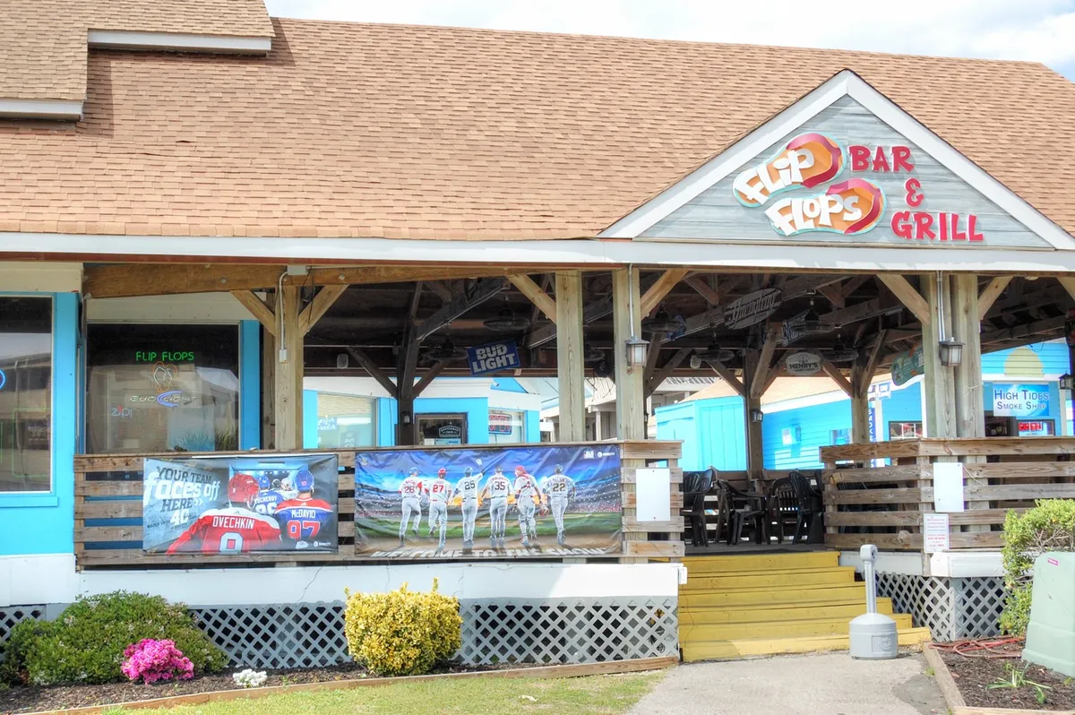 The image shows the exterior of 'Flip Flops Bar & Grill', featuring vibrant signage, colorful banners, and an inviting outdoor seating area. The environment is lively, with people gathered to dine, socialize, and watch sports in a relaxed, open-air atmosphere.