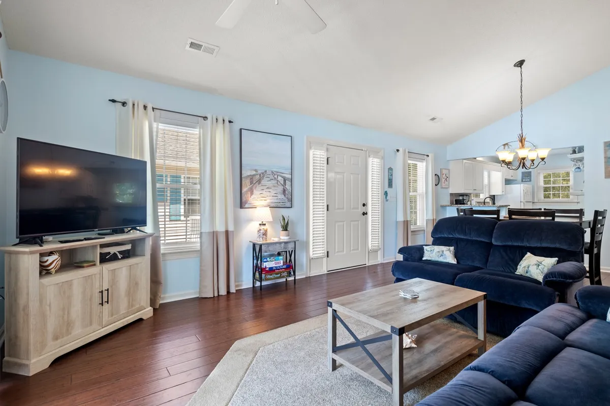 This image captures a cozy, open living and dining area. The layout showcases a TV unit, plush seating, the entrance door, and visible artwork, with the dining table positioned conveniently nearby. The furniture arrangement and lighting create an inviting environment ideal for socializing and family gatherings.
