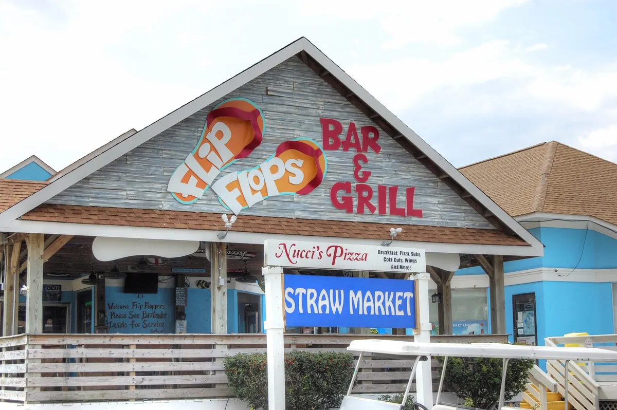This exterior image showcases the lively signage for Flip Flops Bar & Grill, Nucci's Pizza, and the Straw Market, capturing a bustling area filled with dining and shopping options. The scene embodies the fun, social atmosphere that draws both locals and visitors to nearby attractions.