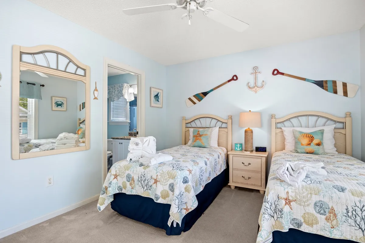 A tranquil bedroom with two twin beds, adorned in nautical-themed bedding. Decorative oars on the wall reinforce the beachside motif, while a mirror reflects the relaxing, sunlit environment. The room is set up to accommodate families or groups needing flexible sleeping arrangements.