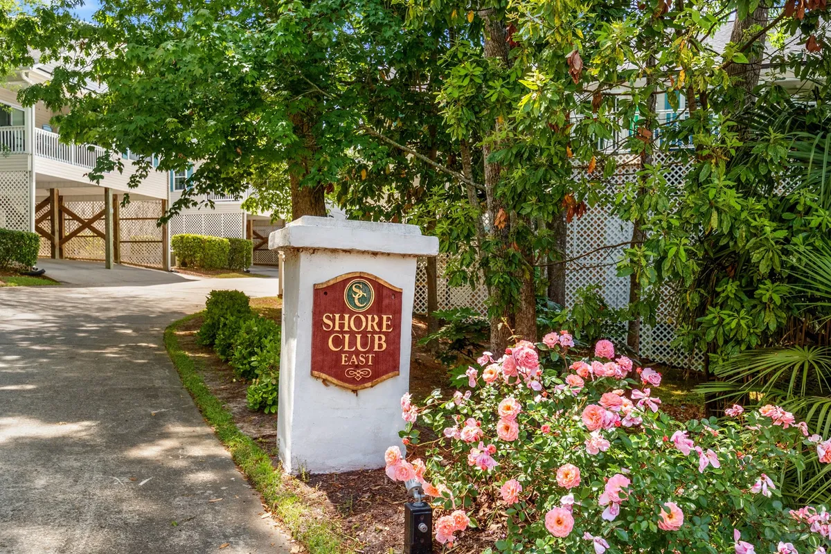 A bright exterior image featuring the Shore Club East sign, bordered by a colorful flower bed and a winding pathway. The scene exudes a sense of arrival and community, with greenery and floral displays framing the entry point to the area.