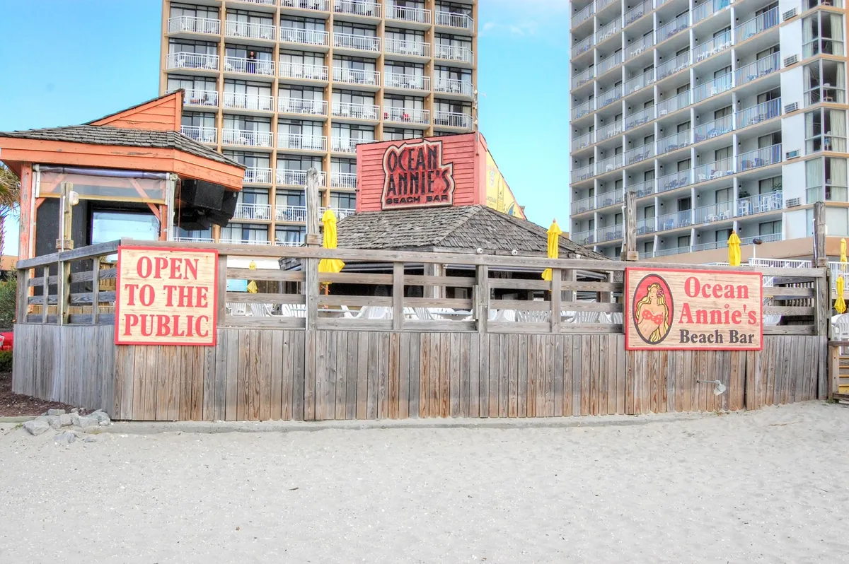 This exterior image captures the beachside setting of Ocean Annie's Beach Bar, with its welcoming sign, wooden fencing, rows of beach chairs under umbrellas, and an 'Open to the public' banner. The scene suggests relaxation and enjoyment right by the ocean.