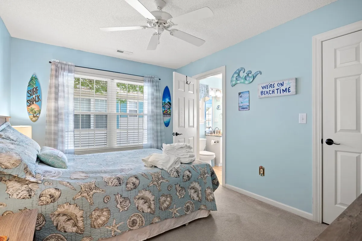 A bright bedroom featuring a King-sized bed with seashell-patterned bedding, surf-inspired wall art, and a large window with sheer curtains letting in natural daylight. The space feels airy and restful, evoking a coastal retreat ideal for relaxation after lively days.