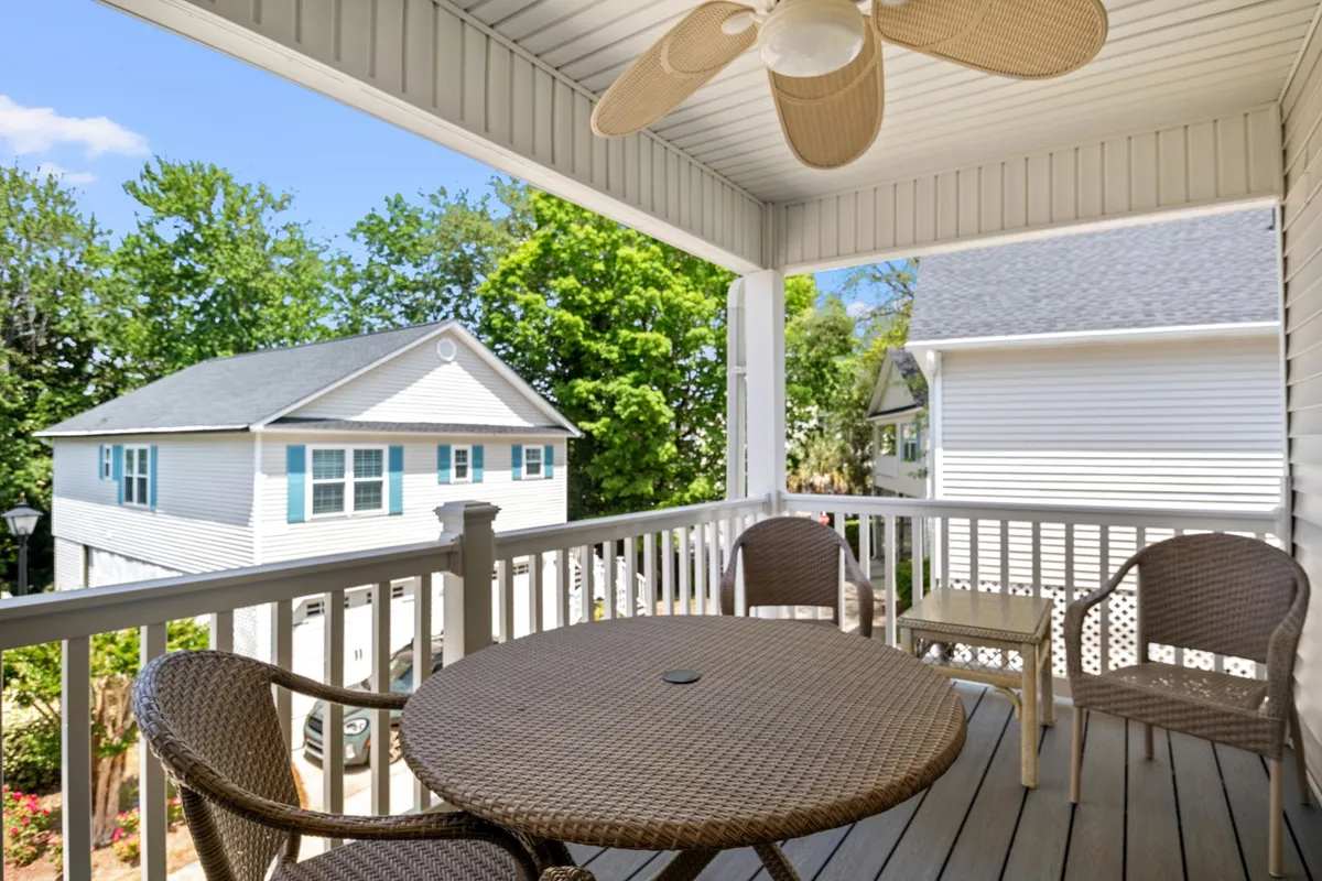The image features the exterior of a raised beach cottage, focusing on a breezy porch furnished with a table and chairs, a ceiling fan overhead, and lush greenery surrounding the space. The setting conveys a serene, shaded spot ideal for unwinding, sharing meals, or simply enjoying the coastal ambiance.