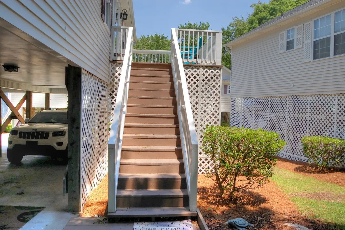 This exterior shot captures the stairs leading up to the cottage's deck, with inviting lattice work, lush bushes, and tasteful landscaping. The upper deck provides a vantage point for outdoor gatherings and relaxation, representing the cottage's welcoming and flexible outdoor spaces.
