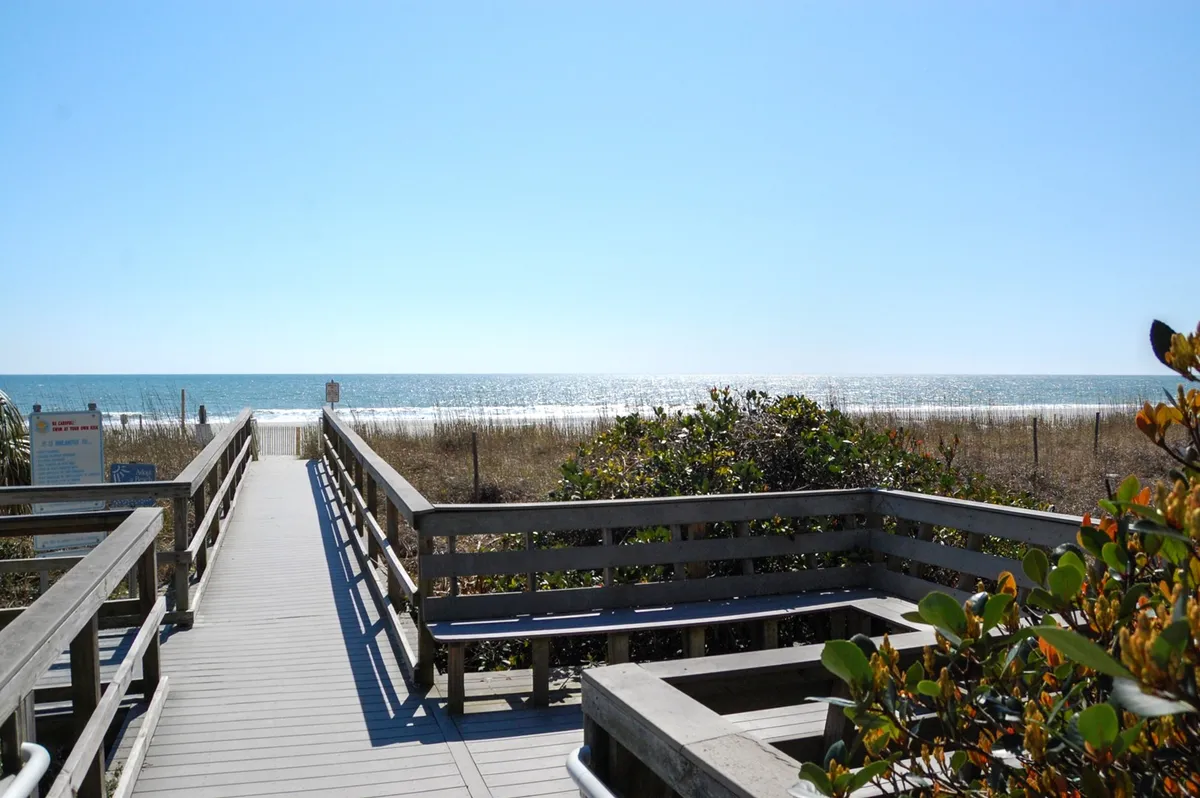 A serene view of a wooden boardwalk leading out over dune grass toward the ocean, offering a glimpse of the beach just moments away. The natural landscaping and shoreline create a sense of proximity and easy access to the coast.