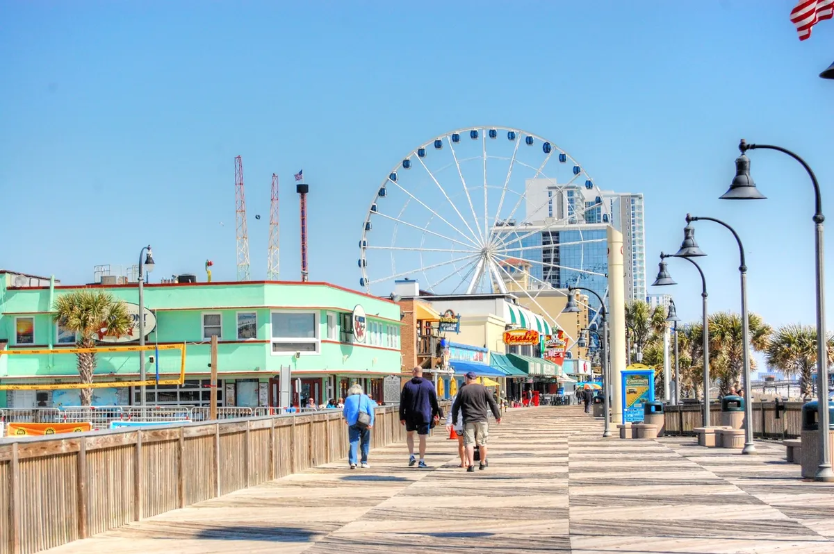 A lively scene of the Myrtle Beach Boardwalk featuring the iconic ferris wheel (SkyWheel), colorful oceanfront buildings, and people enjoying walks and local attractions. The photo captures the vibrant, festive atmosphere of the boardwalk area.