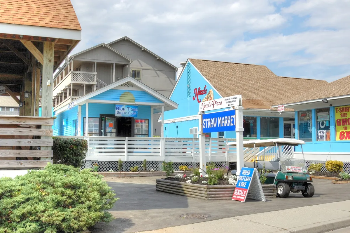 Depicting the colorful exteriors of local shops, the Straw Market, High Tide Smoke Shop, and Nino's Pizza, this lively scene teems with activity, inviting guests to explore, shop, and savor local specialties.